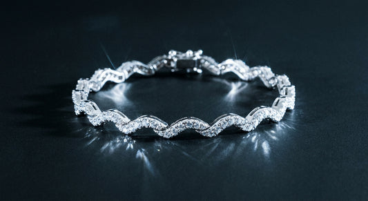 A silver wave tennis bracelet adorned with sparkling stones photographed against a dark matte surface with dramatic icy light reflections.